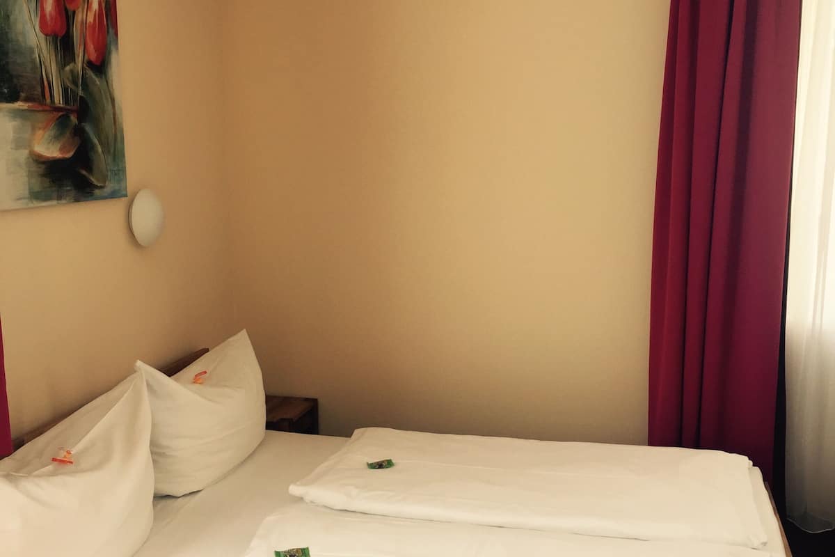 A hotel room with a wooden bed frame, white bedding, a painting of flowers on the wall, and a red curtain.