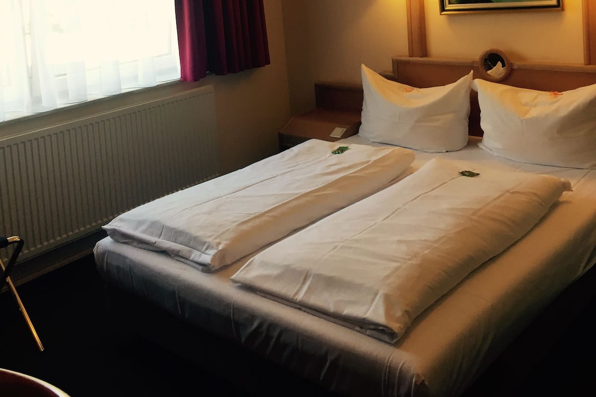 A hotel room with a bed, a window with curtains, a painting on the wall, and two bedside lamps.