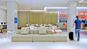 Een moderne hotellobby met een grote beige bank, een televisie en een barruimte met rode decoratie.