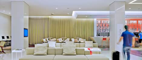 A modern hotel lobby with a large beige sofa, a TV, and a bar area with red decor.