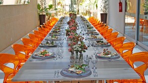 Une longue table à manger, entourée de chaises orange, est dressée avec des assiettes, des verres et des couverts.