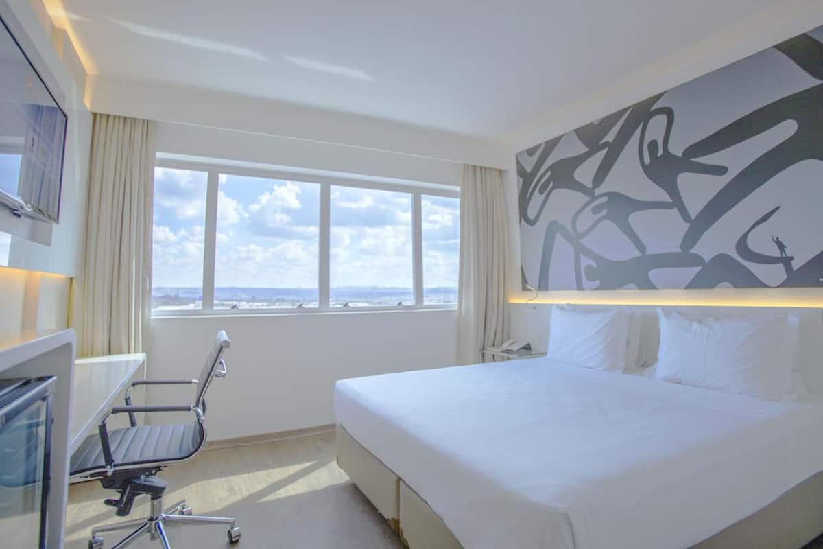 Una habitación de hotel con cama, escritorio, silla, un amplio ventanal con vistas y una obra de arte en la pared.