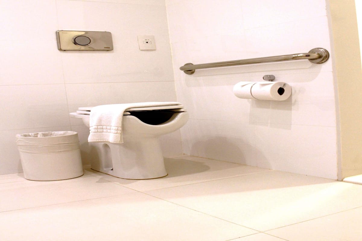 Un baño con inodoro, un bote de basura, un dispensador de papel higiénico y un botón de descarga en la pared.