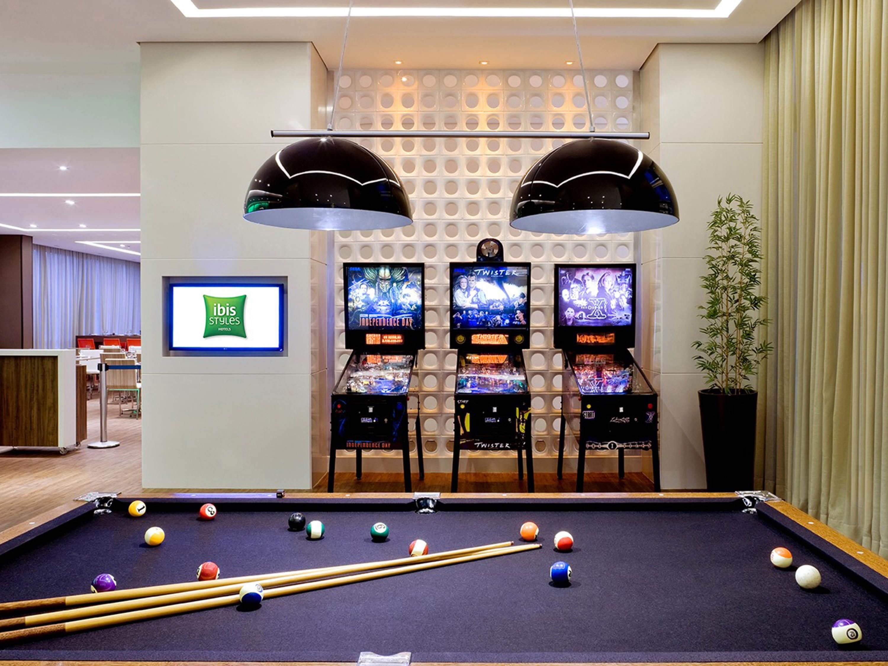 A pool table with balls and cues, three arcade machines, and two overhead lamps in a room with a white wall and a green sign.