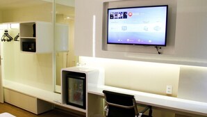 Een moderne hotelkamer met een flatscreen televisie aan de muur, een kleine koelkast, een bureau met een stoel en een nachtkastje.