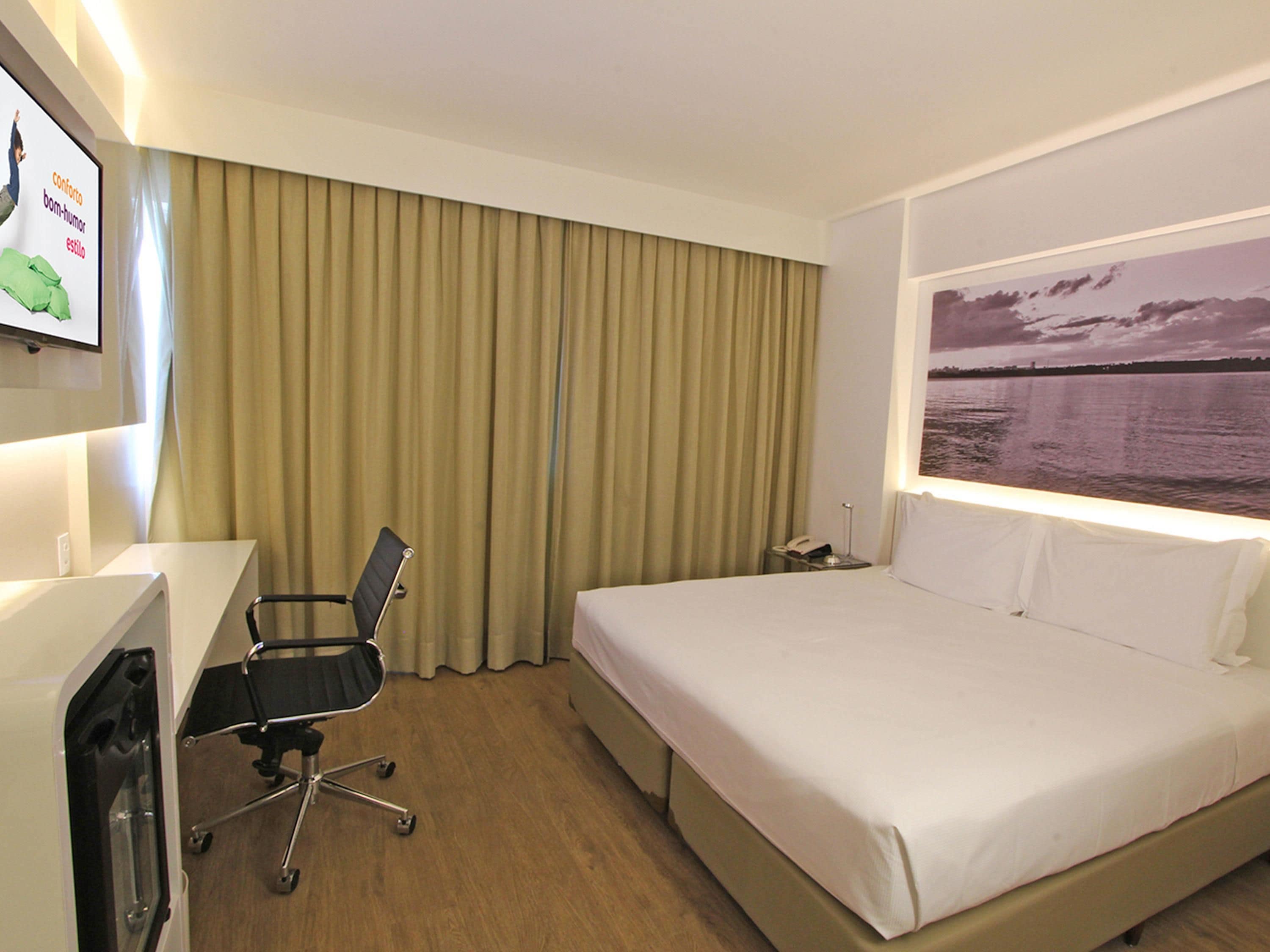 A hotel room with a bed, a desk, a chair, a phone, and a painting of a sunset over water.