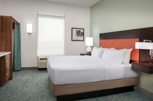 Habitación de hotel con una cama grande, una mesita de noche con lámpara, un armario de madera, una ventana con persianas y un cuadro en la pared.