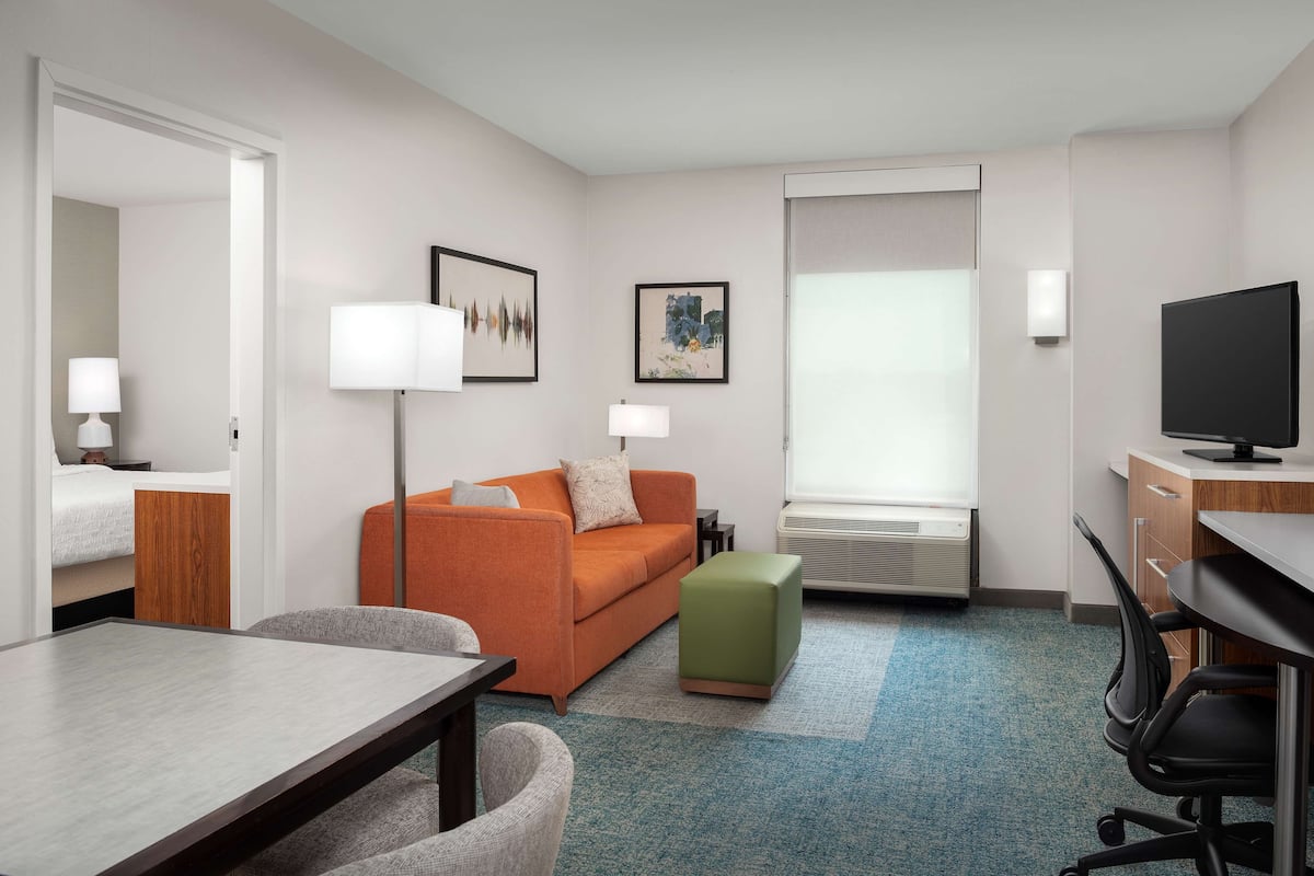 Habitación de hotel con cama, sofá, escritorio, televisor y ventana con aire acondicionado.