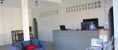 Zona de recepción con un mostrador gris, un sofá morado con un cojín rojo, y una mesa con revistas y una computadora portátil.