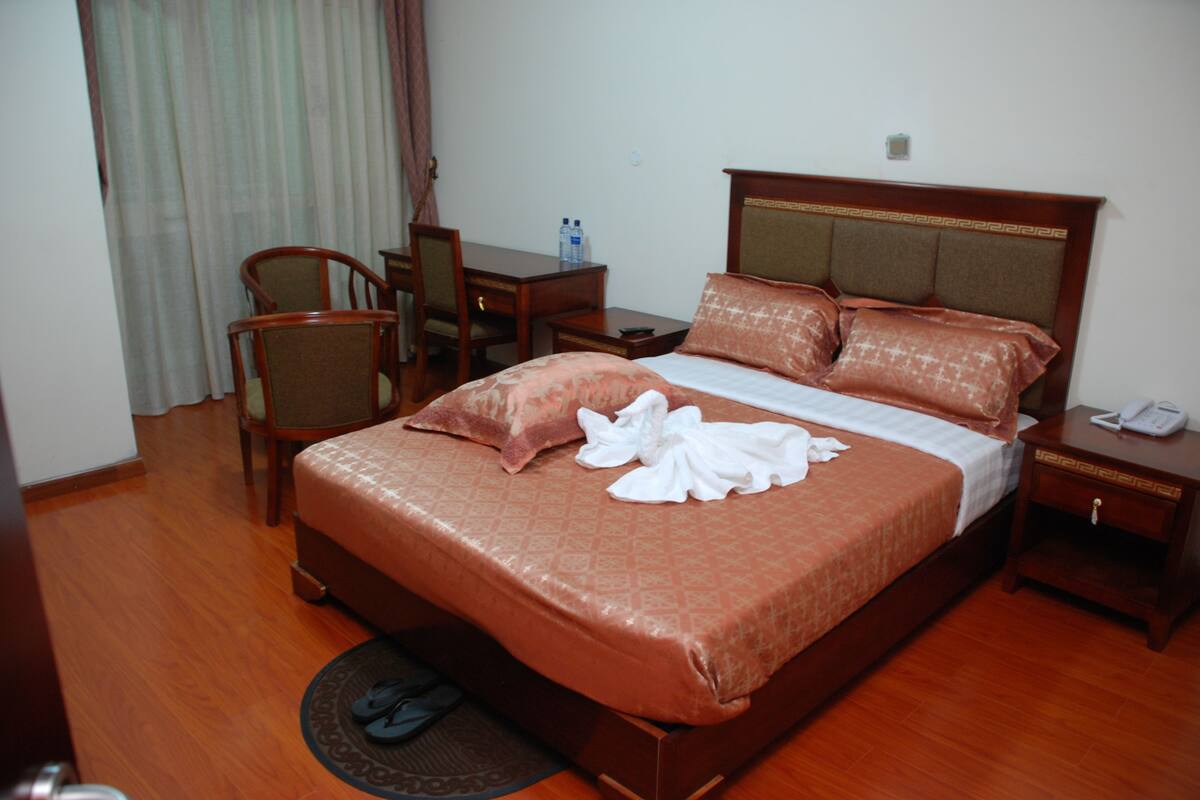 A hotel room with a wooden bed, a desk with a chair, a telephone, and a water bottle on the nightstand.