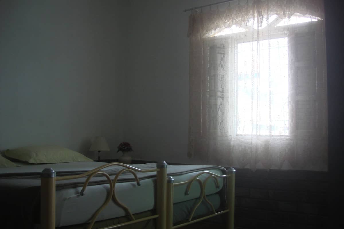 A dimly lit room with a single bed, a small table with a lamp, and a window with sheer curtains.