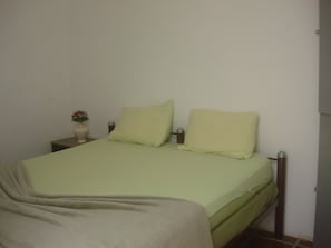 Un lit simple avec une couvre-lit verte et des oreillers, une table de chevet ornée d’un vase de fleurs et un mur uni.