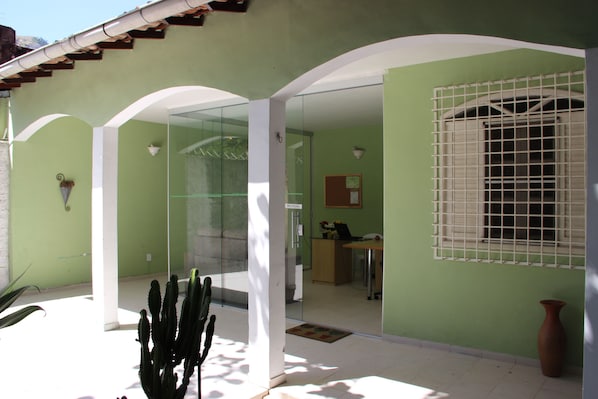 Un bâtiment à l’extérieur verdoyant, doté de colonnes blanches et d’une porte en verre donnant sur un espace intérieur où se trouvent un bureau et une chaise.