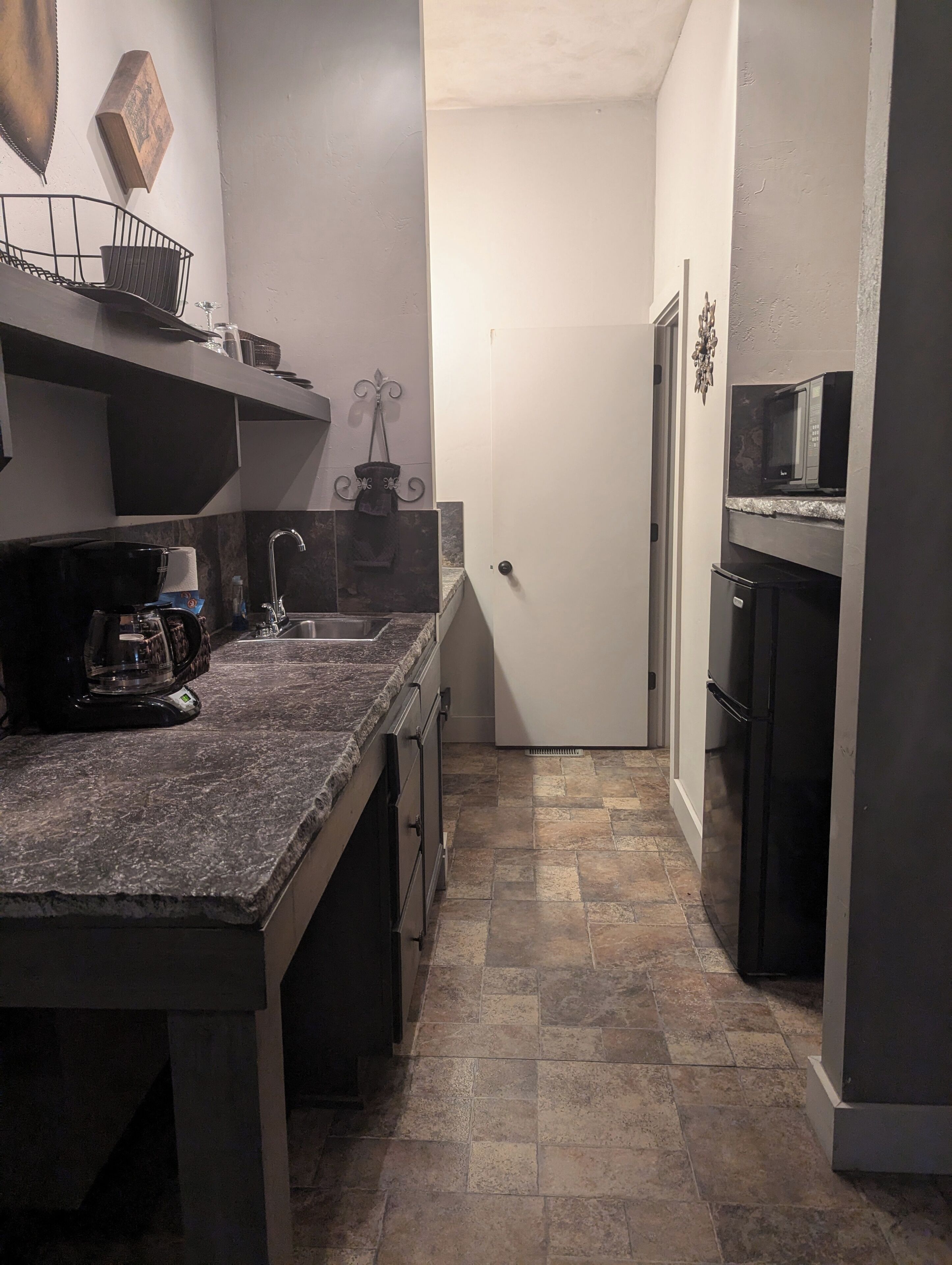 A narrow kitchen with a granite countertop, a black coffee maker, a microwave, and a refrigerator.