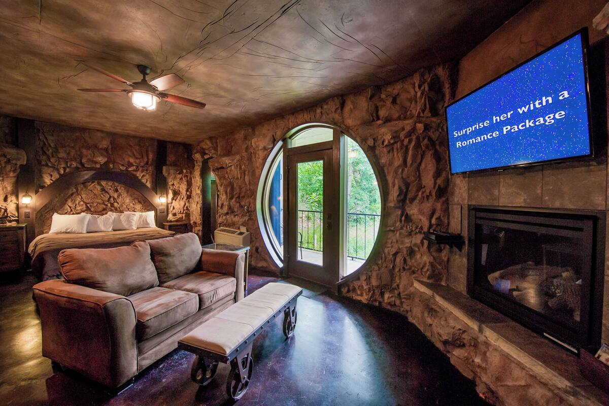 Una habitación acogedora con pared de piedra, chimenea, cama, sofá y televisor que muestra el texto: 'Sorpréndela con un Paquete Romántico'.