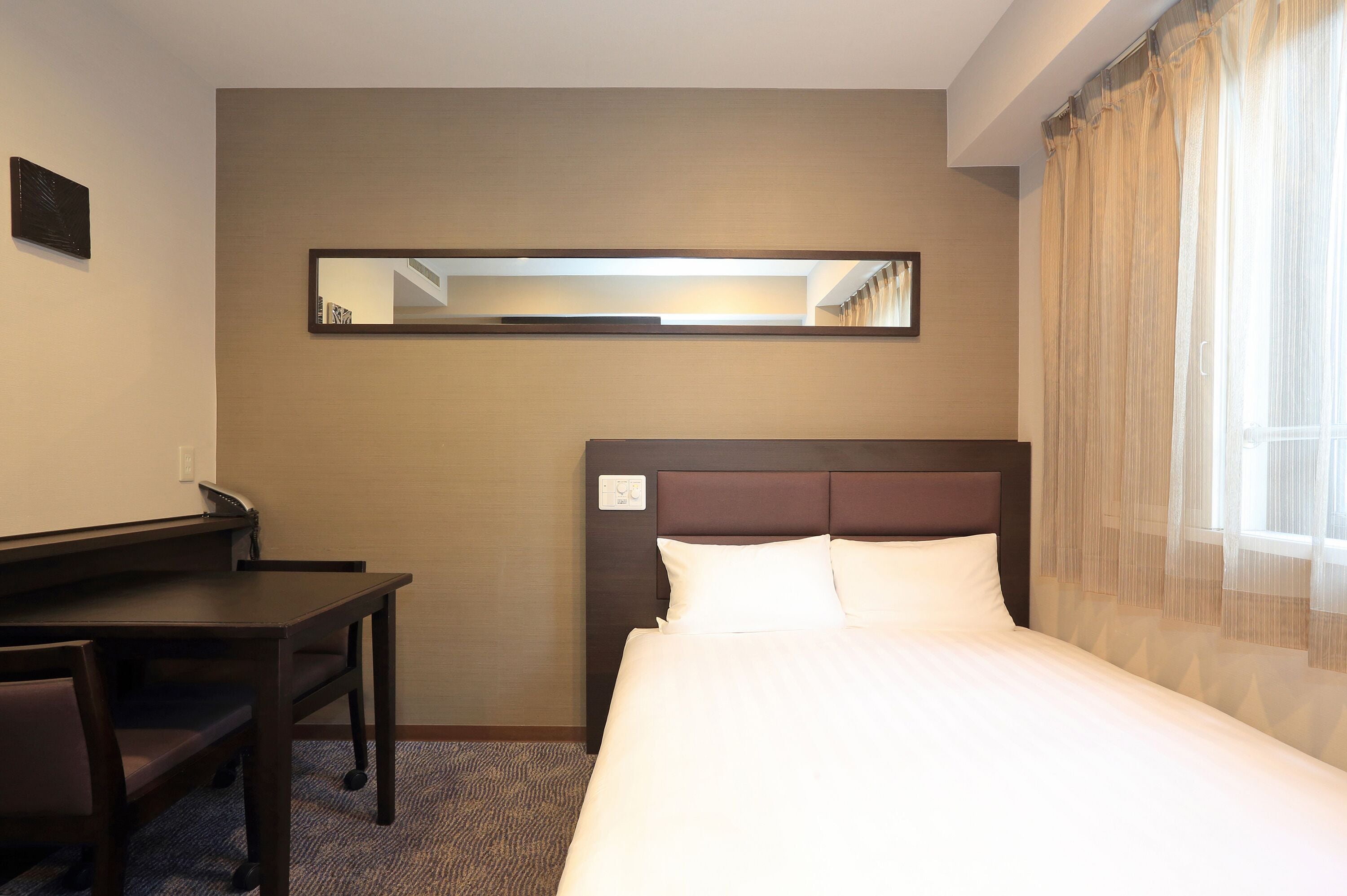 A hotel room with a bed, a desk, and a chair. There is a mirror on the wall and a window with curtains.