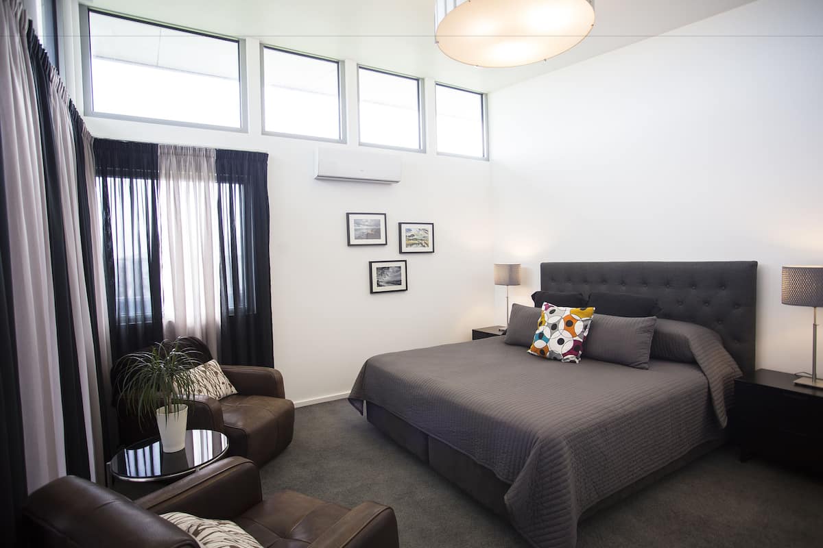 A modern hotel room with a large bed, a sitting area with a sofa and armchair, a small table with a plant, and a large window with curtains.