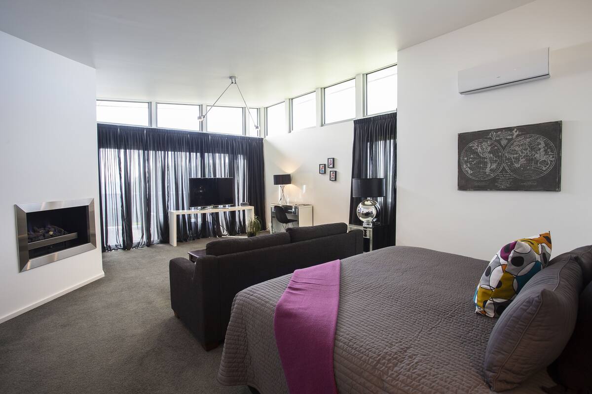 A modern hotel room with a large bed, a sofa, a TV, and a fireplace.
