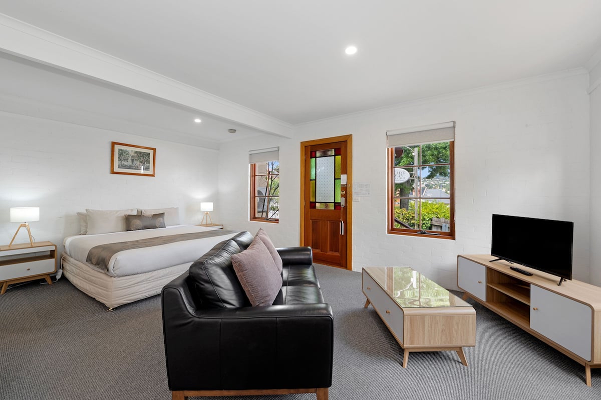 A modern hotel room with a bed, a black leather sofa, a TV, and a glass coffee table.