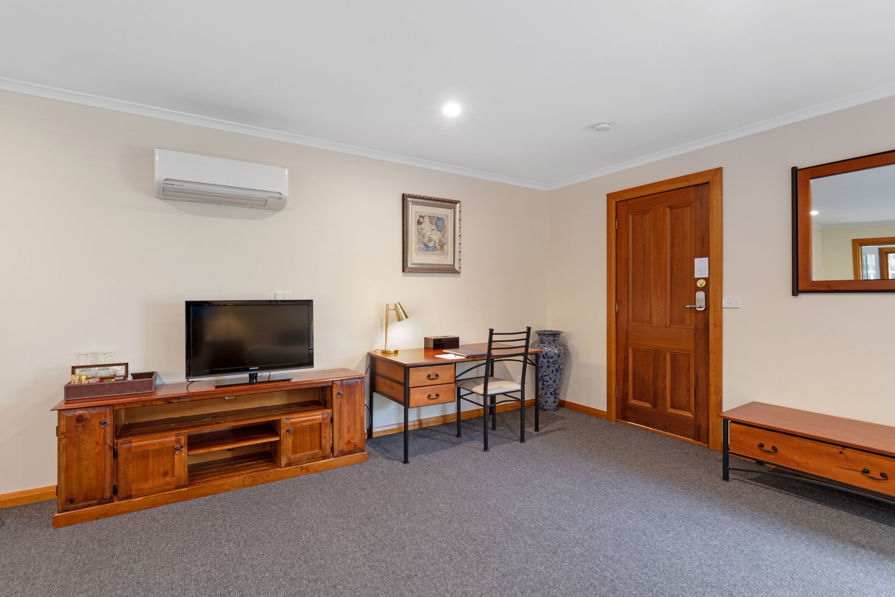 A room with a television on a wooden entertainment unit, a desk with a chair, a wooden door, and a large mirror on the wall.