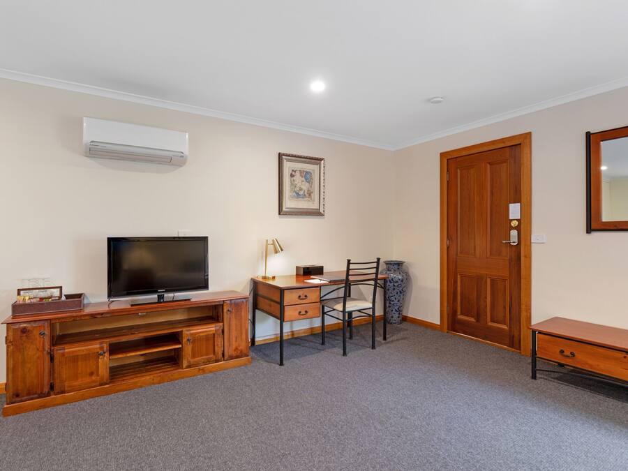 A room with a television on a wooden entertainment unit, a desk with a chair, a wooden door, and a large mirror on the wall.