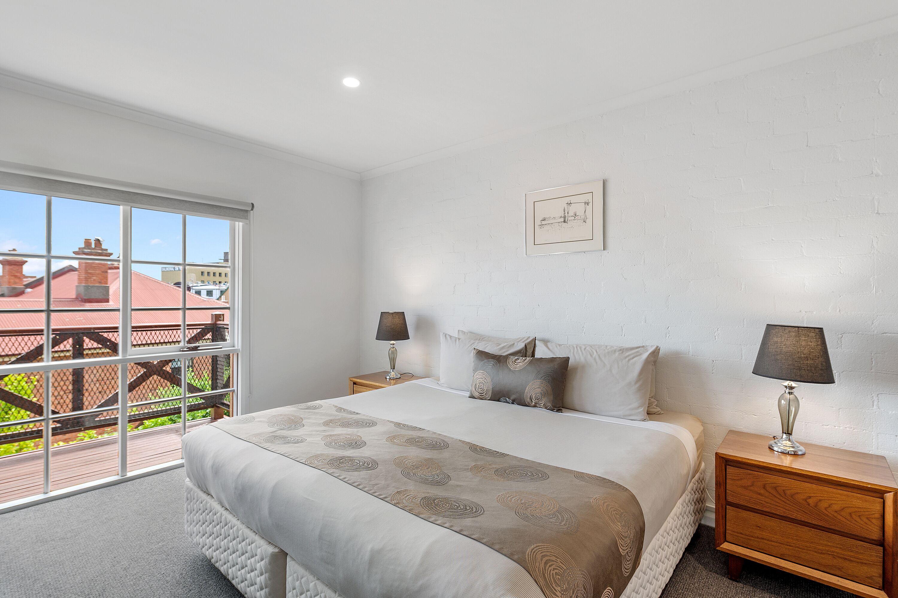 A bedroom with a large window, a bed with a patterned bedspread, two bedside tables with lamps, and a framed picture on the wall.