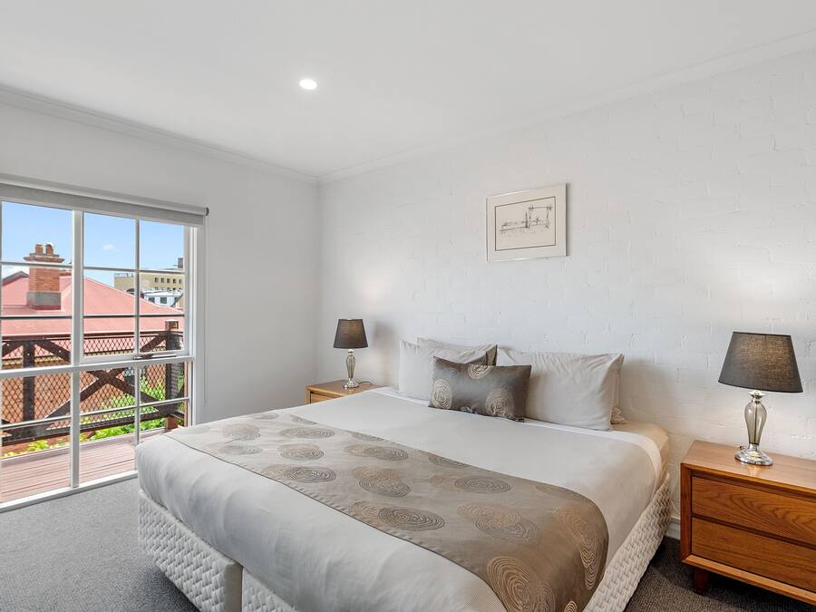 A bedroom with a large window, a bed with a patterned bedspread, two bedside tables with lamps, and a framed picture on the wall.