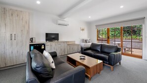 A modern living room with a sofa, coffee table, and television. There is a kitchen area with cabinets and a refrigerator. Large windows provide a view of greenery outside.
