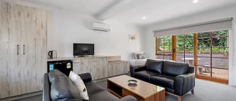 A modern living room with a sofa, coffee table, and television. There is a kitchen area with cabinets and a refrigerator. Large windows provide a view of greenery outside.