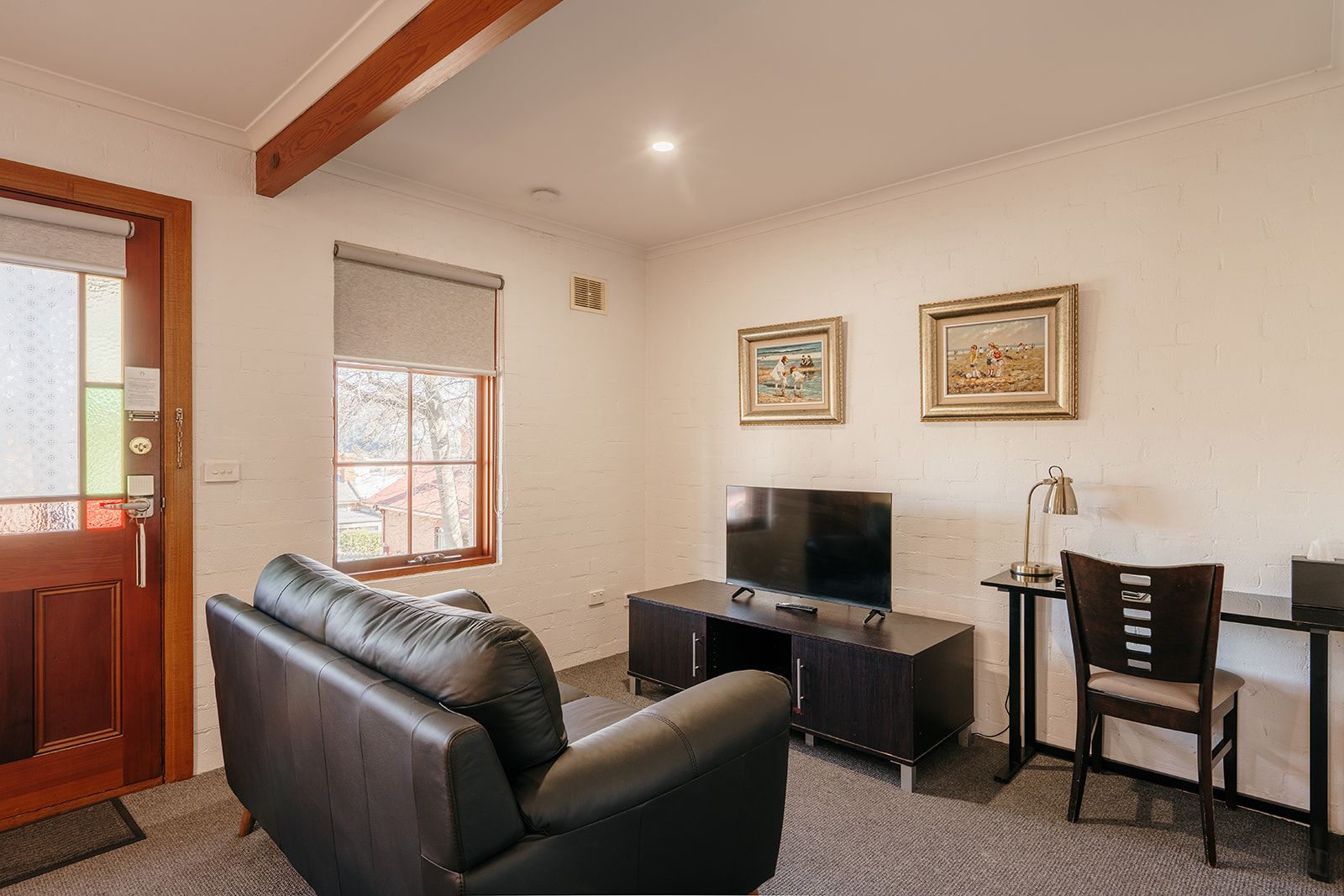 A room with a leather sofa, a TV on a stand, a desk with a chair, and two framed pictures on the wall.