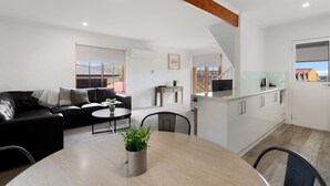 A modern apartment with a living room, dining area, and kitchen. There is a black leather sofa, a round dining table with a potted plant, and a kitchen with white cabinets and a wooden ceiling beam.