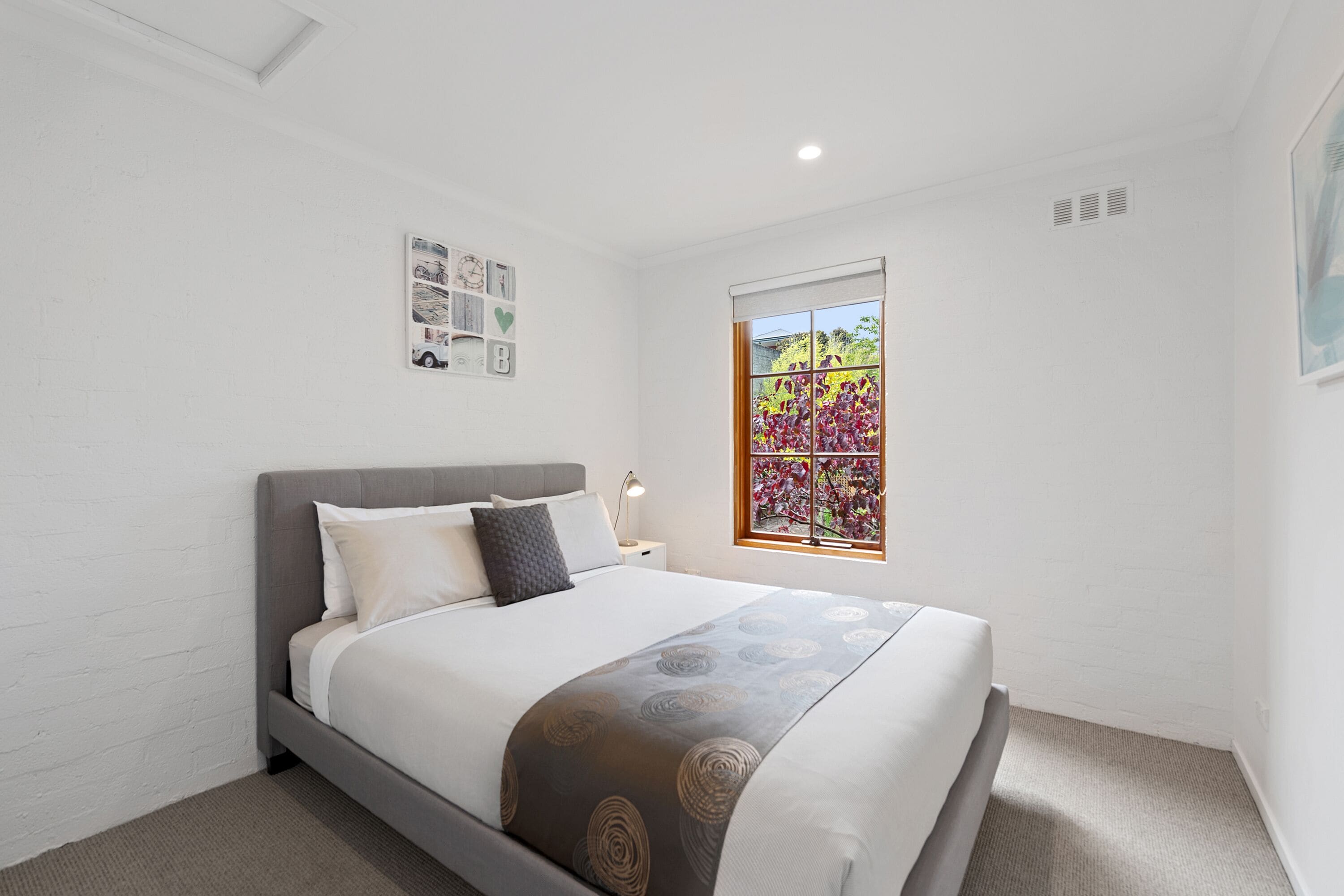 Un dormitorio ordenado con una cama, una ventana que da a flores, y una pared con un reloj y una decoración con forma de corazón.
