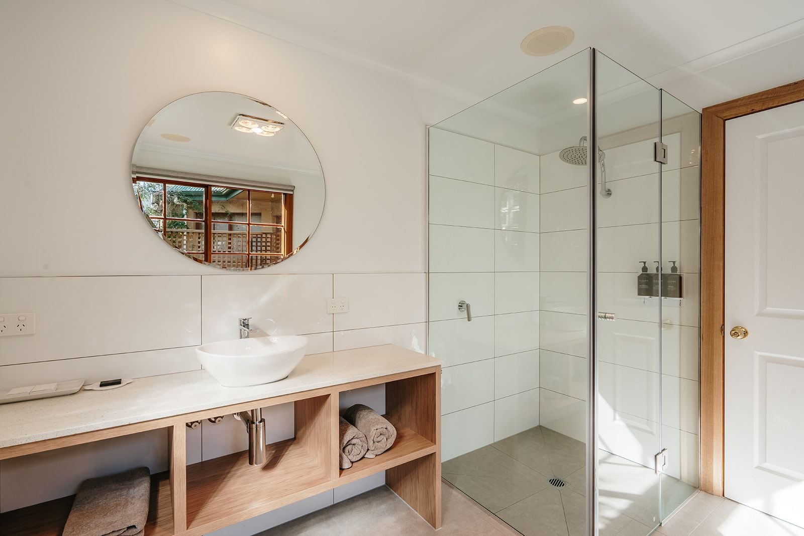 Une salle de bain moderne avec un grand miroir rond, un lavabo blanc, une douche en verre et un meuble-lavabo en bois.