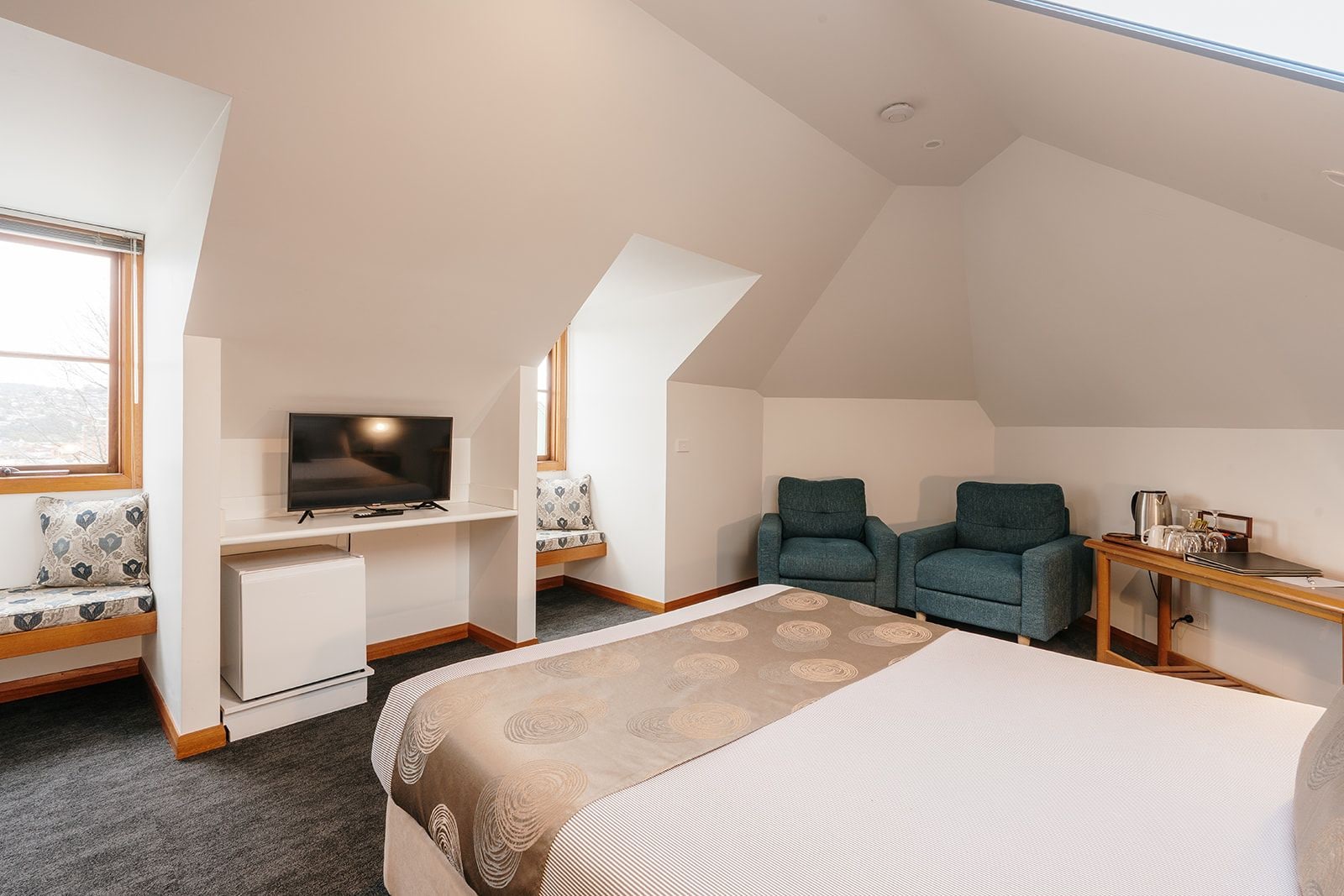 Habitación de hotel moderna con cama, escritorio con televisor, dos sillones y una mesita con hervidor.