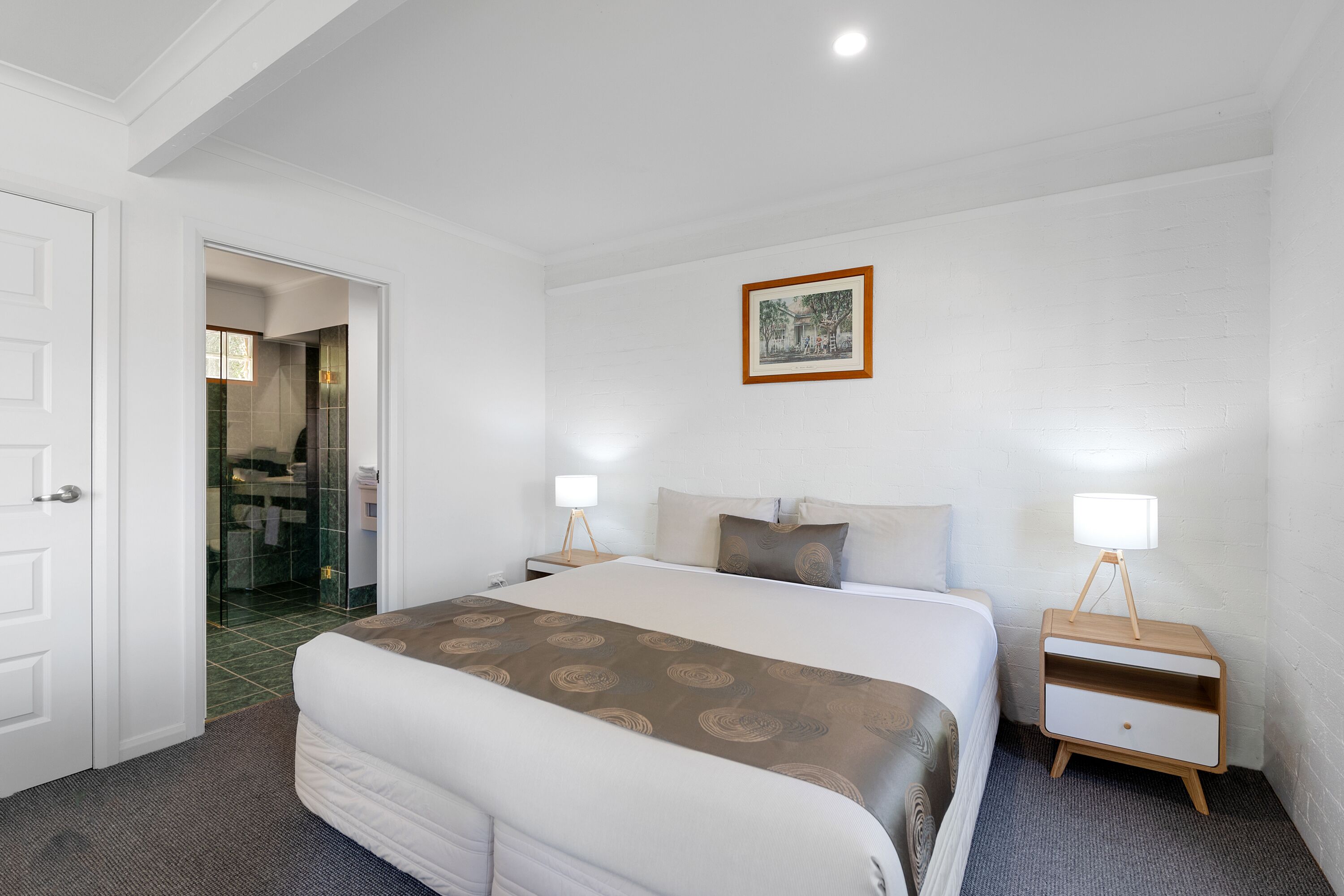 Ein Hotelzimmer mit einem großen Bett, zwei Nachttischen, einem kleinen Tischchen, einem gerahmten Bild an der Wand und einem durch eine offene Tür einsehbaren Badezimmer.