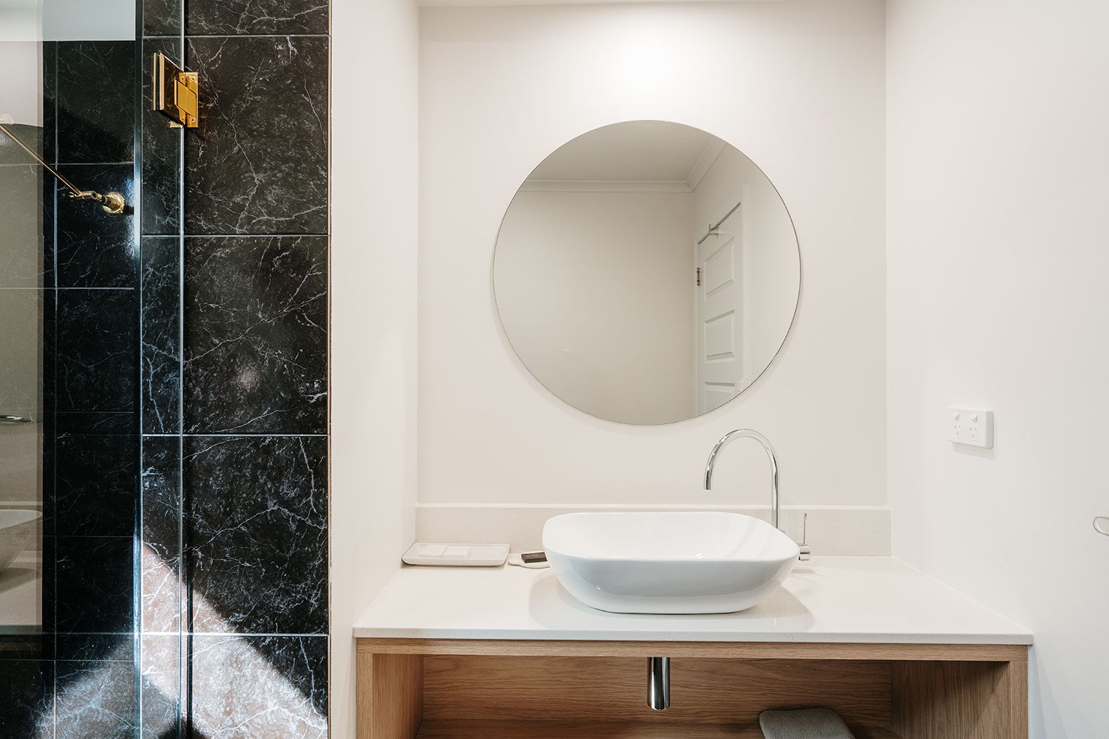 Un baño moderno con tocador blanco, espejo redondo y cabina de ducha de mármol negro.