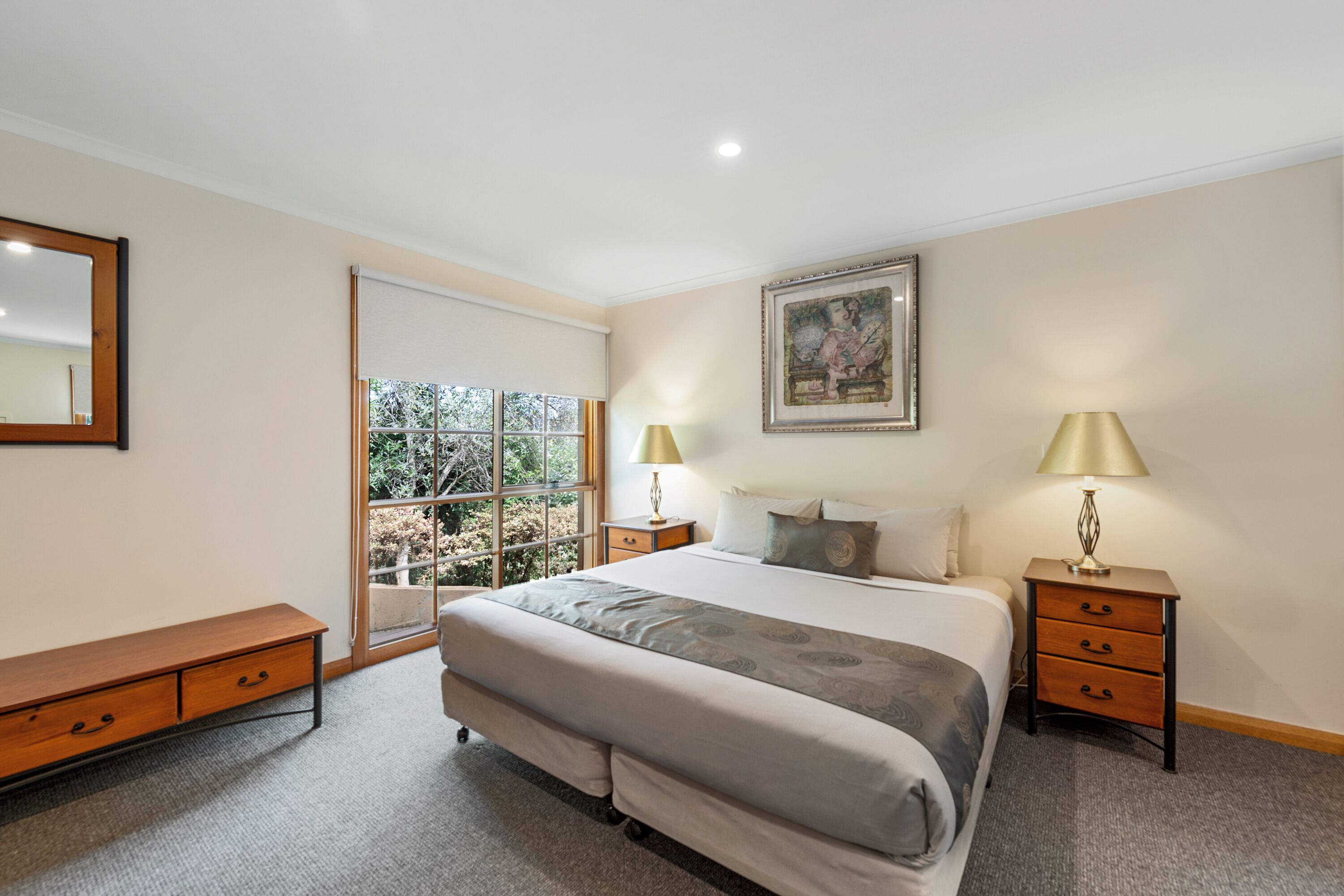 A bedroom with a large bed, two bedside tables with lamps, a bench, a window with a view, and a framed picture on the wall.