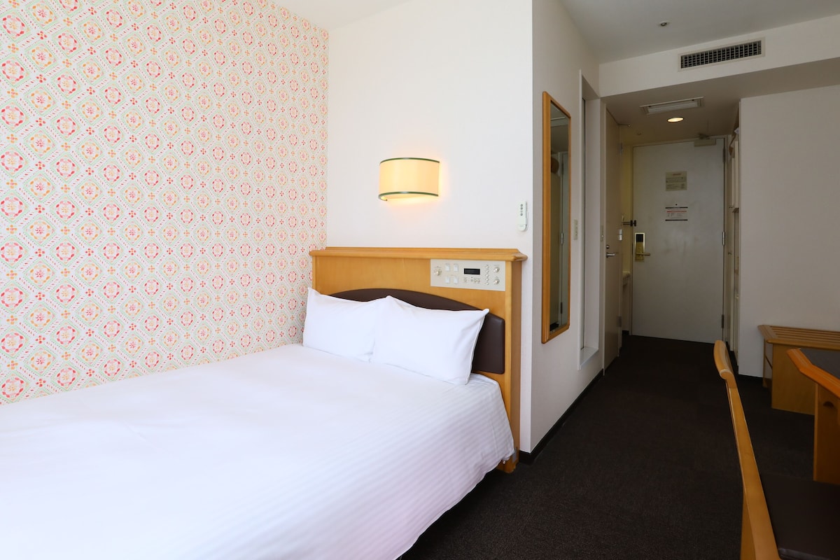 Ein Hotelzimmer mit einem Bett, einer an der Wand befestigten Steuerung, einem Spiegel und einer Tür zu einem weiteren Raum.