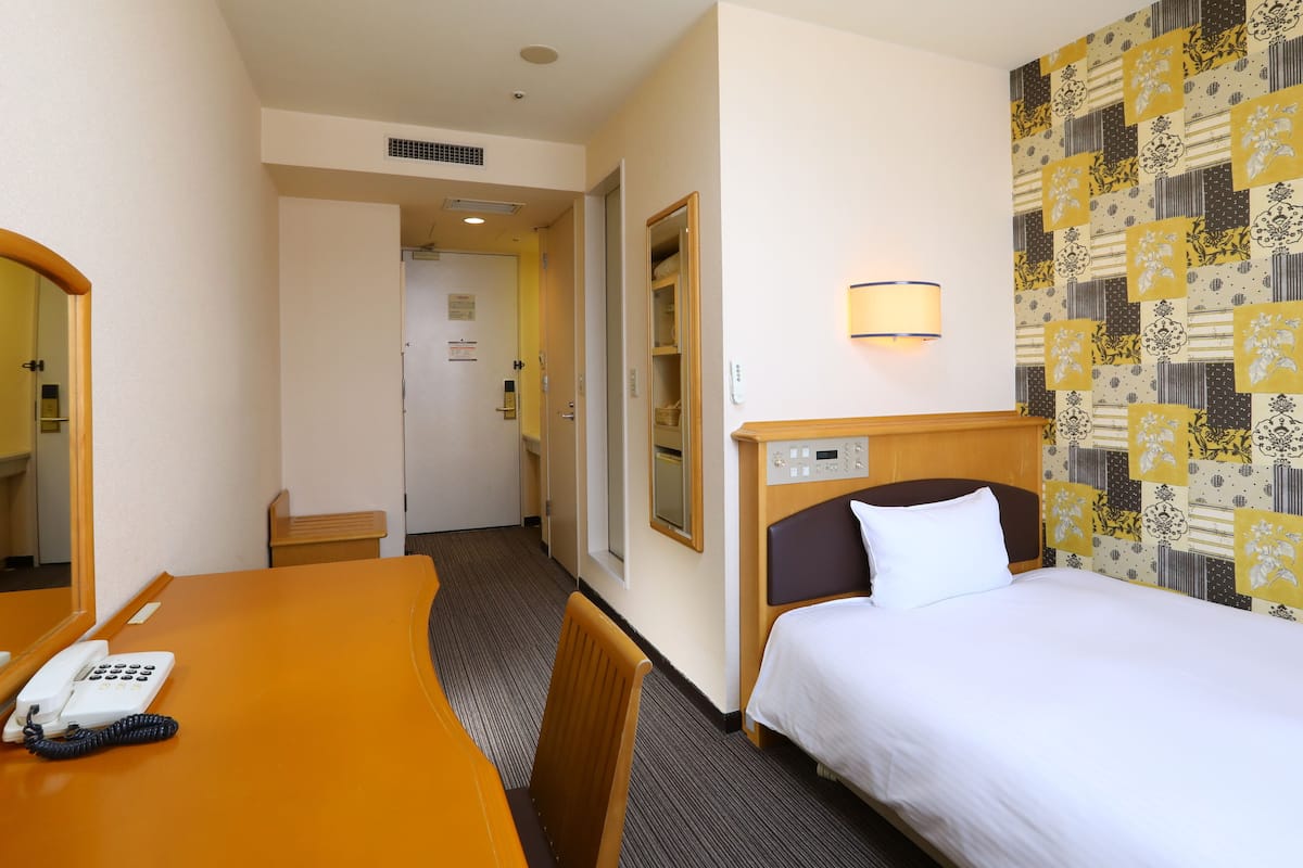 Ein Hotelzimmer mit einem Bett, einem Schreibtisch mit Telefon, einem Spiegel und einer Wand mit ornamentalem Wandpapier.