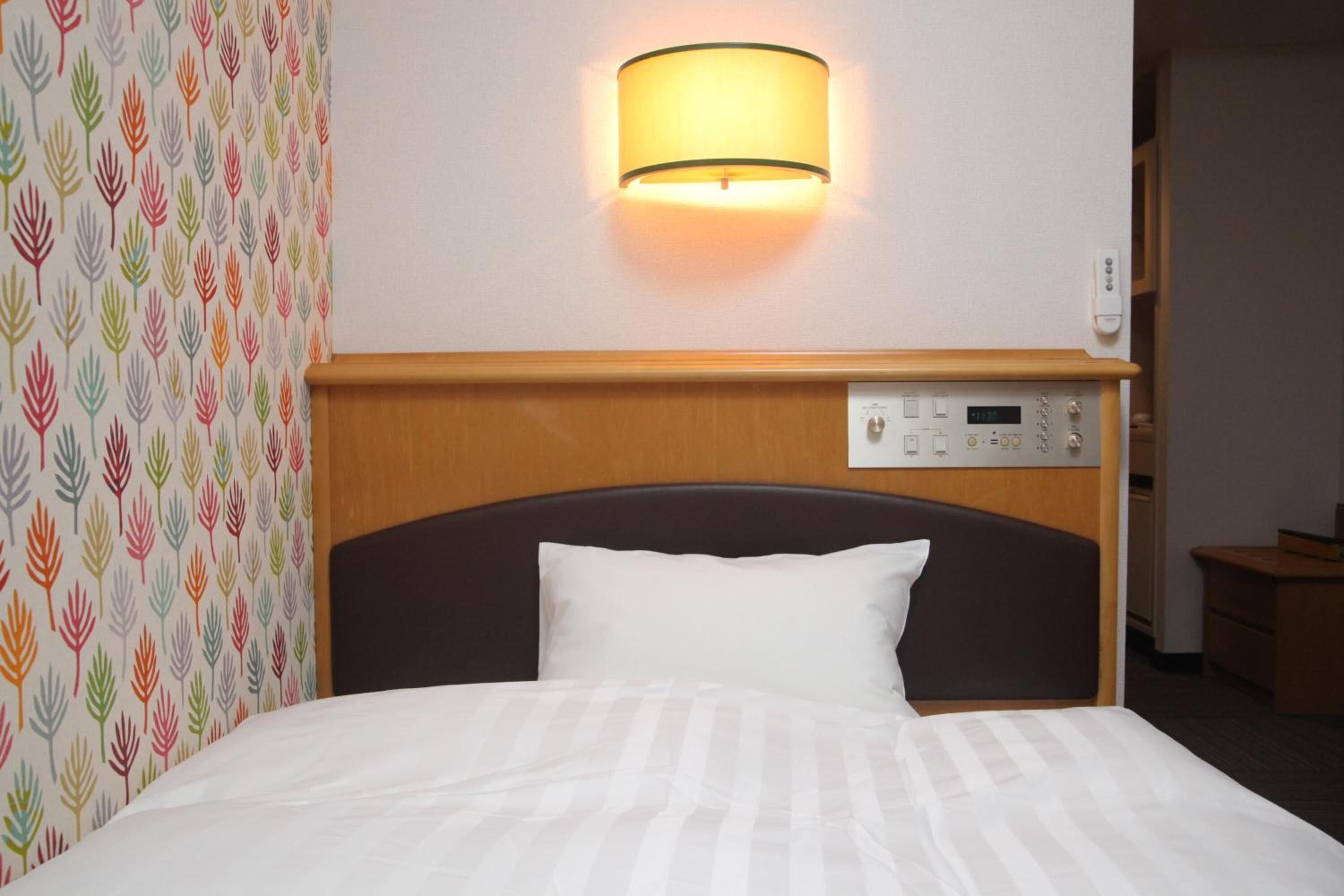 A hotel room with a bed, a wall-mounted control panel, and a patterned wallpaper on the left.