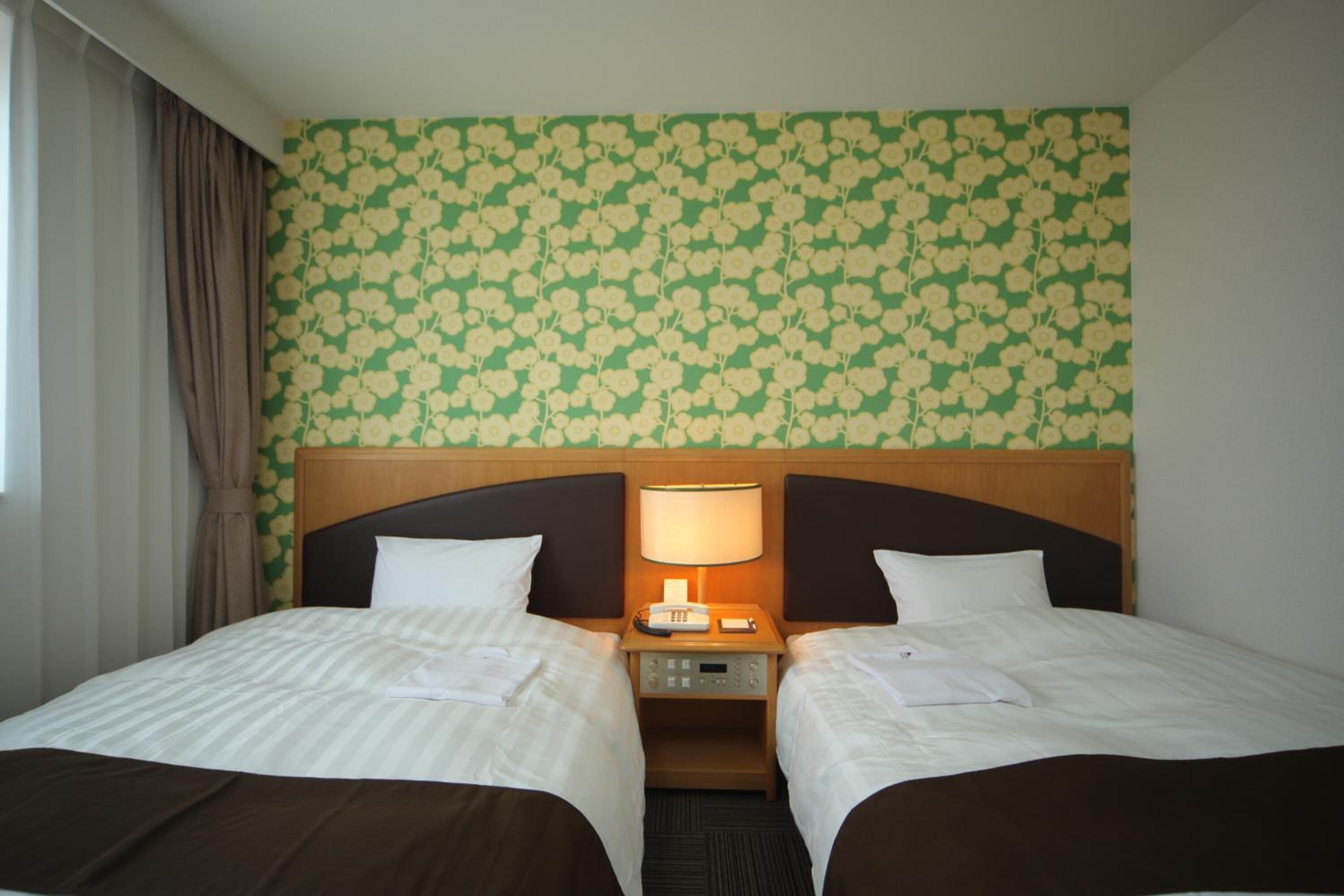 Quarto de hotel com duas camas, uma cabeceira de madeira, uma mesa de cabeceira com abajur e papel de parede floral.