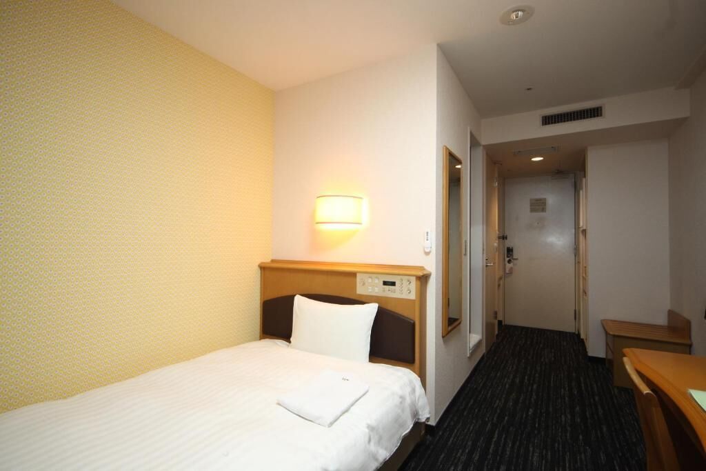 A hotel room with a single bed, a bedside table, a lamp, and a door leading to a bathroom.