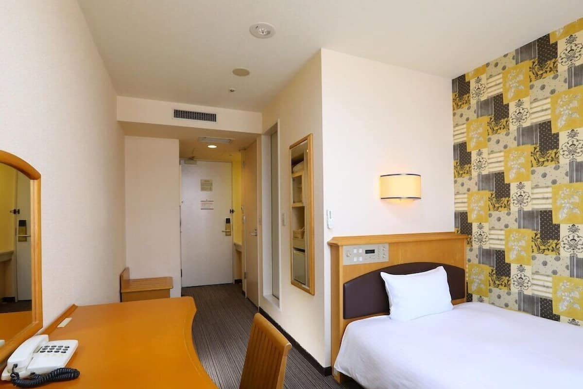 Ein Hotelzimmer mit einem Bett, einem Schreibtisch mit Telefon, einem Spiegel, einer Wandleuchte und einem Wandteppich.