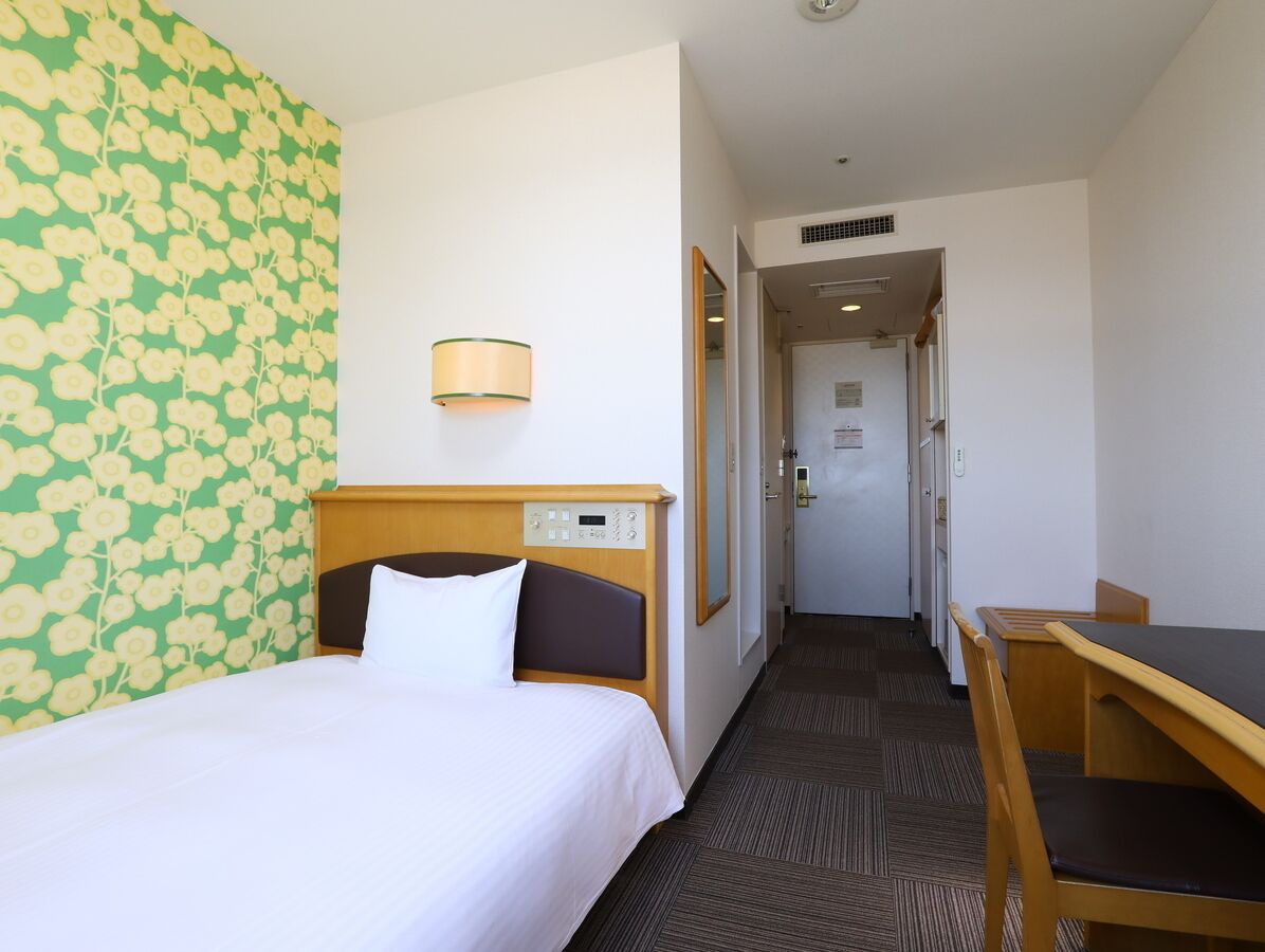 Quarto de hotel com uma cama, uma escrivaninha e uma cadeira. Há uma parede com padrão floral e um abajur na parede.