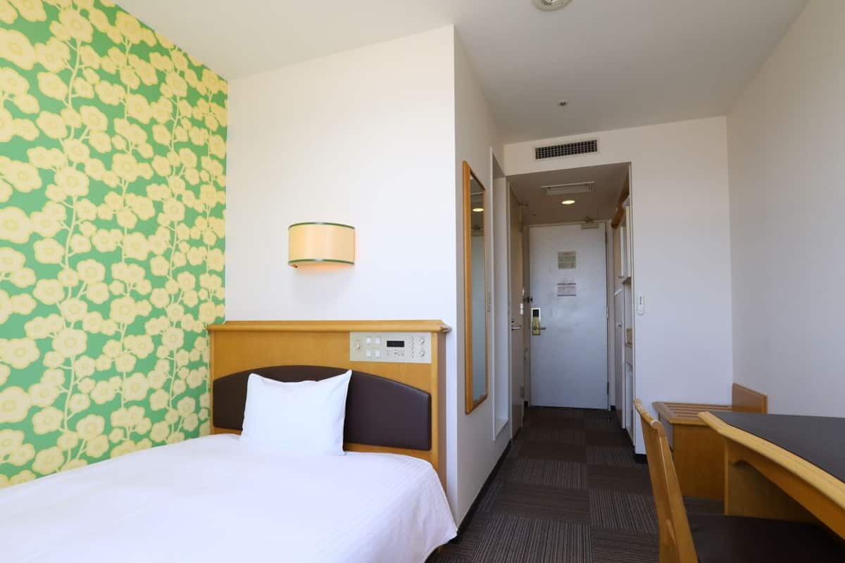 Ein Hotelzimmer mit einem Bett, einem Schreibtisch und einem Stuhl. An der Wand befindet sich ein Blumenmuster und eine Lampe.