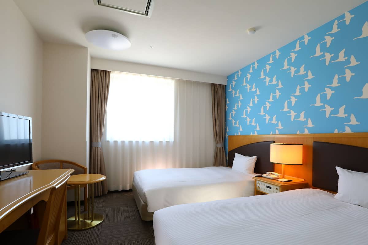 Ein Hotelzimmer mit zwei Betten, einem Schreibtisch, einem Fernseher, einer Lampe und einem Fenster mit Vorhängen.