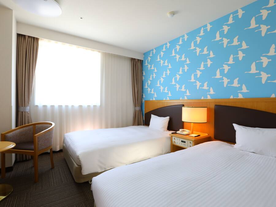 A hotel room with two beds, a chair, a nightstand with a lamp, and a window with curtains.