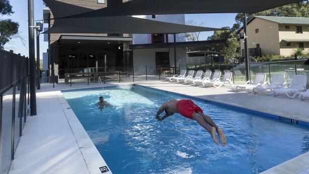 A swimming pool with a person diving, surrounded by lounge chairs and a building in the background. - ECU Joondalup Village (Joondalup)