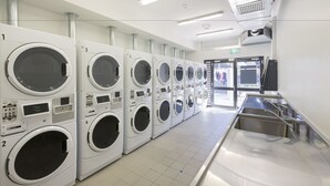 A row of white front-loading washers with control panels, a stainless steel sink, and a door leading outside. - ECU Joondalup Village (Joondalup)