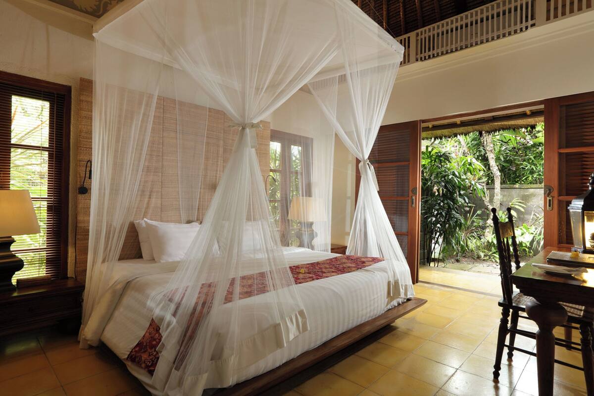A room with a four-poster bed, a wooden table, and a view of greenery outside.